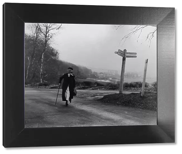 Elderly nurse walking down a country lane past a crossroad sign, c1930