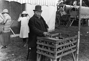 Showman in front of coconut shy, horse-drawn caravans in background, 1920s